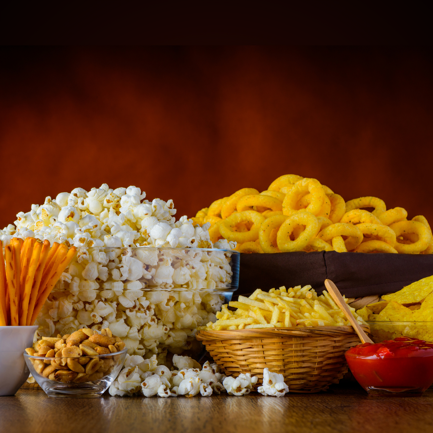 An assortment of snacks including popcorn, fries in a basket, onion rings, pretzel sticks, nachos, and nuts, all displayed on a wooden surface with a dark background.
